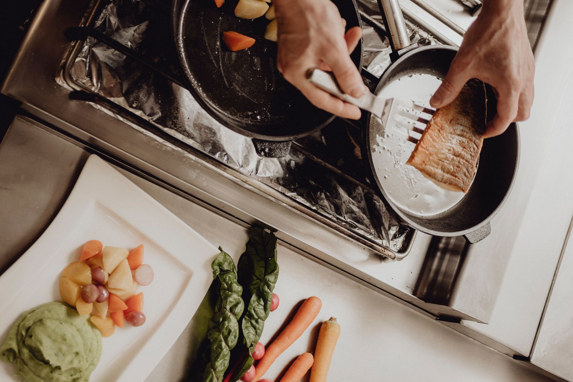 Chef prepares salmon in a pan, vegetables and puree on a plate next to it.