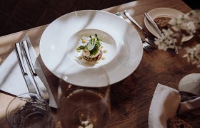 A plate of smoked trout tartare on a potato pancake, garnished with herbs and flowers, on a wooden table with cutlery and a glass of wine.
