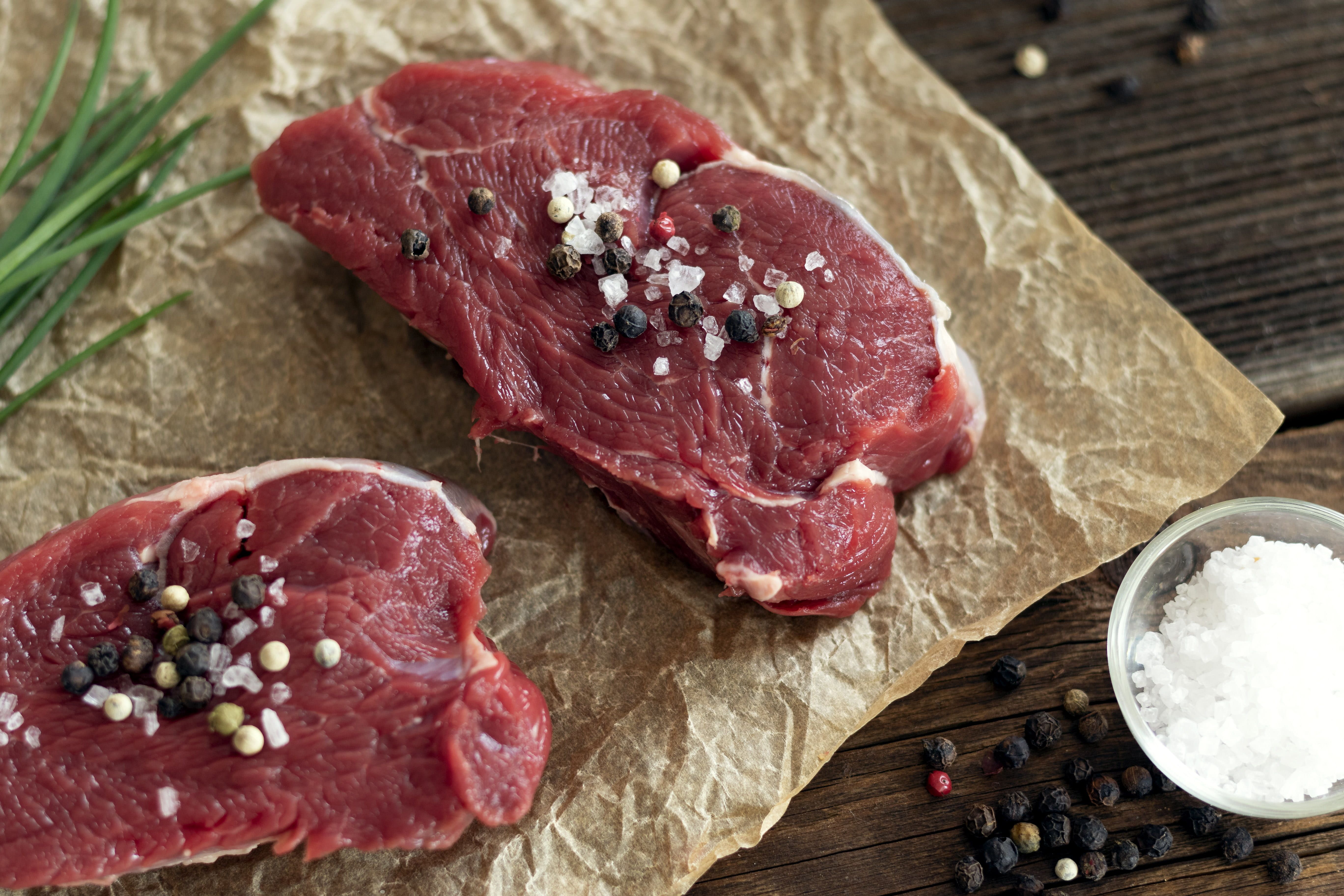 Two pieces of raw beef sprinkled with salt and pepper on baking paper, next to chives, a bowl of salt and pepper.