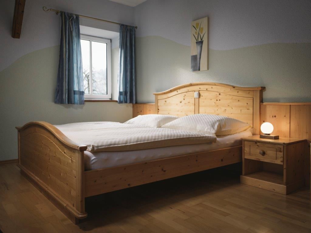 A bedroom with a wooden bed, bedside table and lamp, blue curtains and a picture on the wall.