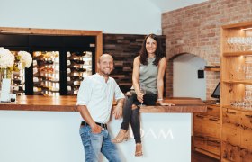 Two people in a modern wine bar, a woman sitting on the counter, a man leaning against it. Wine racks and glasses in the background.