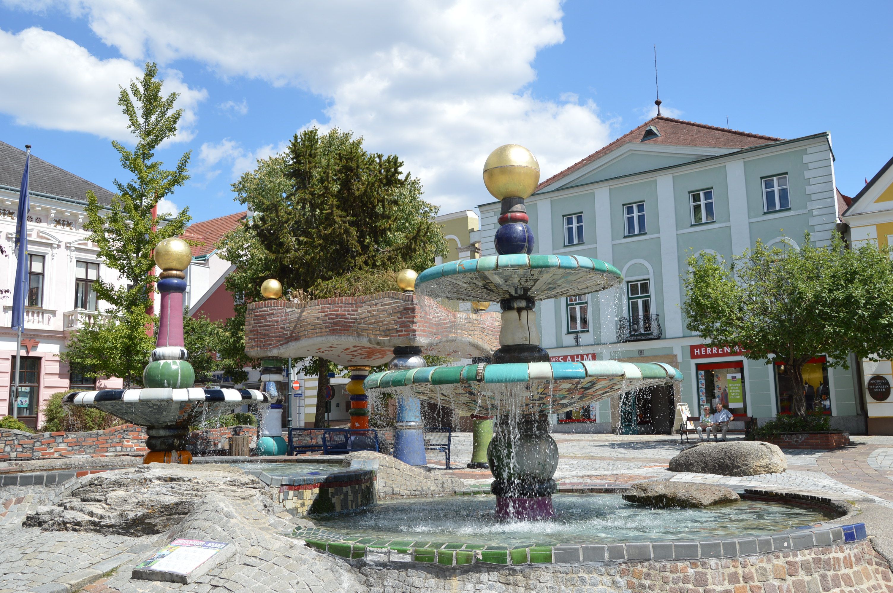 A colorful Hundertwasser-style fountain with golden balls and flowing water, surrounded by buildings and trees.