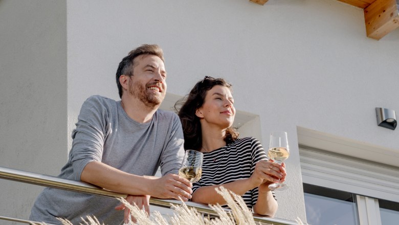 A couple stands on a balcony holding glasses of wine, with a modern building in the background.