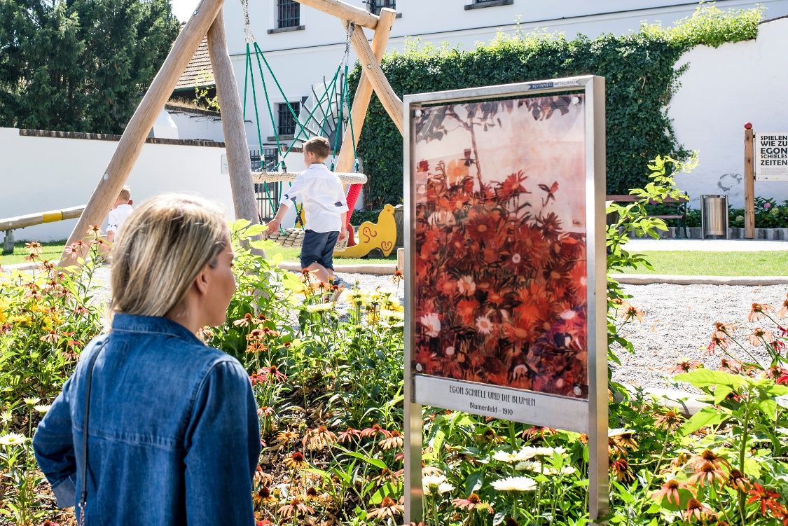A woman looks at a painting by Egon Schiele in a garden with a playground in the background.