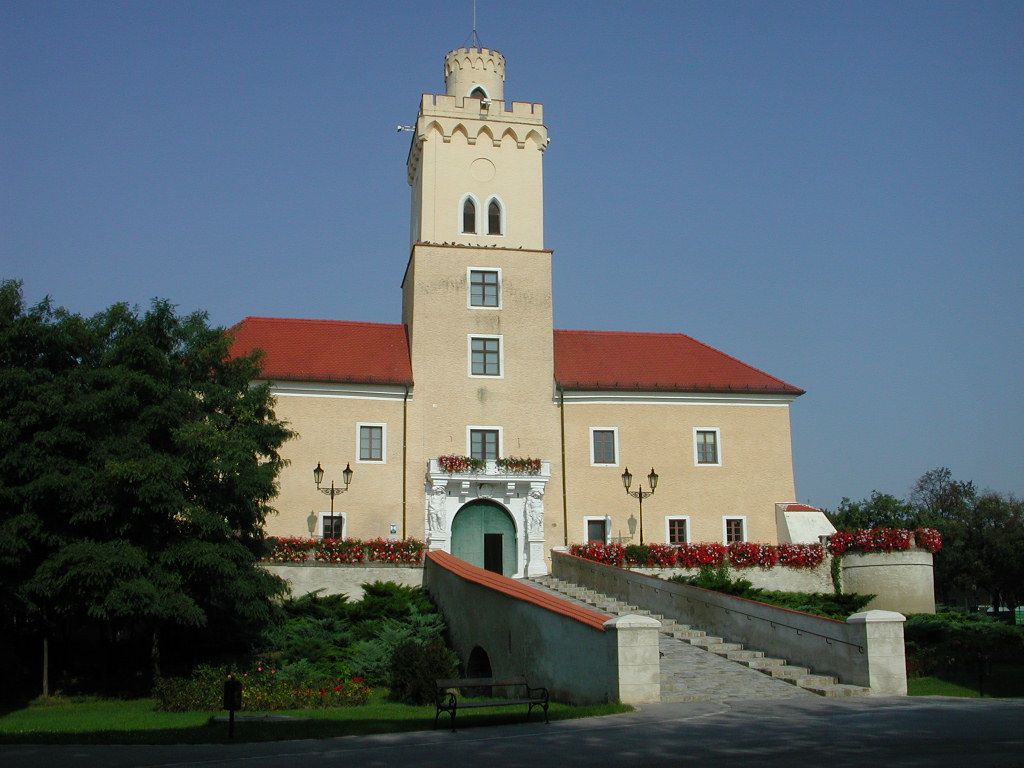 A castle with a tower and red roofs, surrounded by flowers and trees, under a clear blue sky.