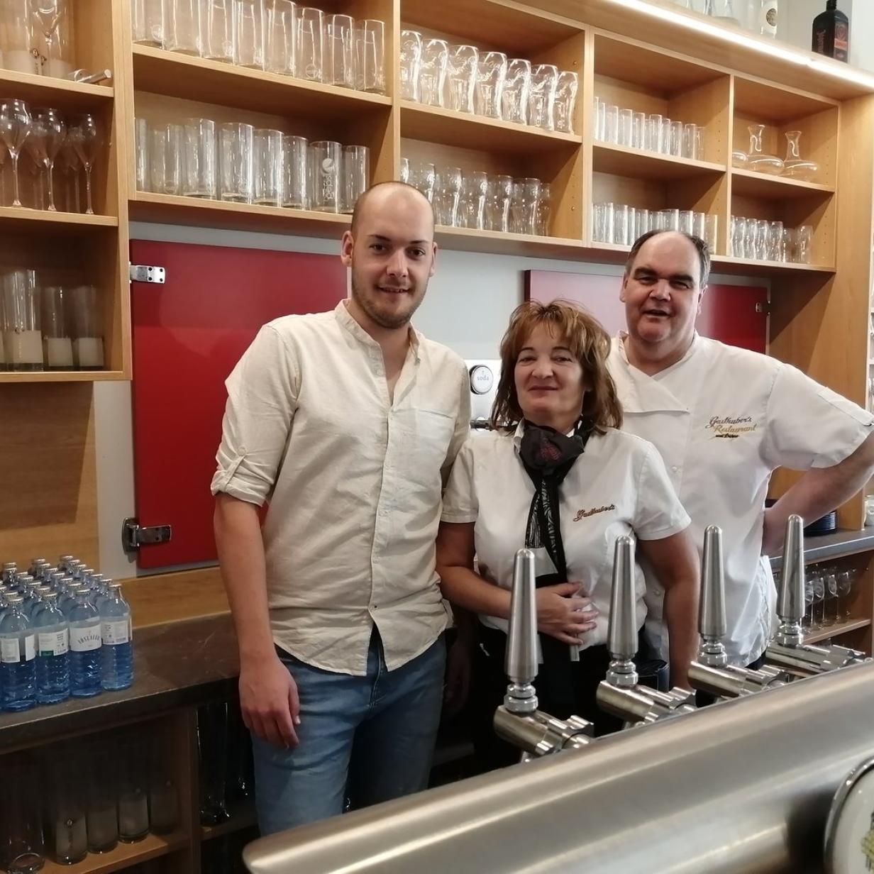 Three people stand behind a bar with taps and glasses in the background.