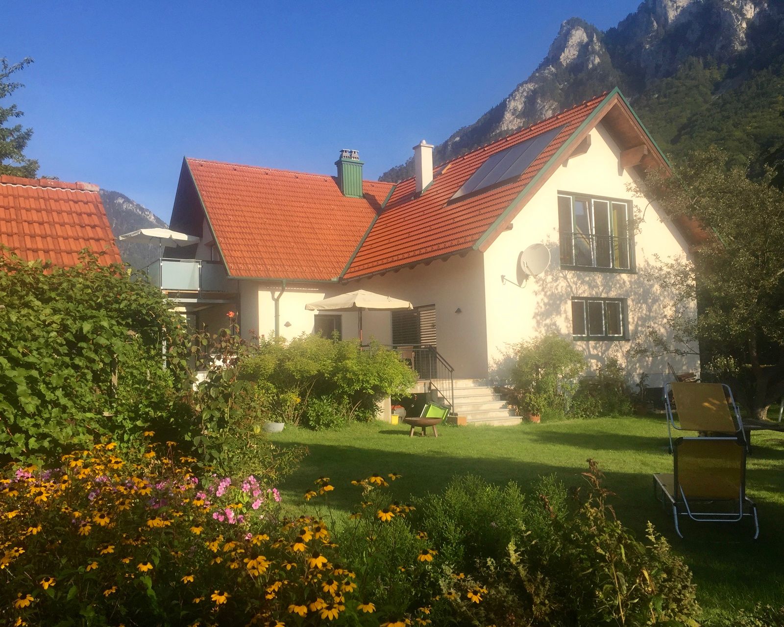 A house with a red roof, surrounded by a garden with flowers and deckchairs, against a mountain backdrop.