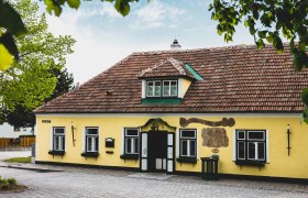Yellow building with red roof tiles and green shutters, surrounded by trees.