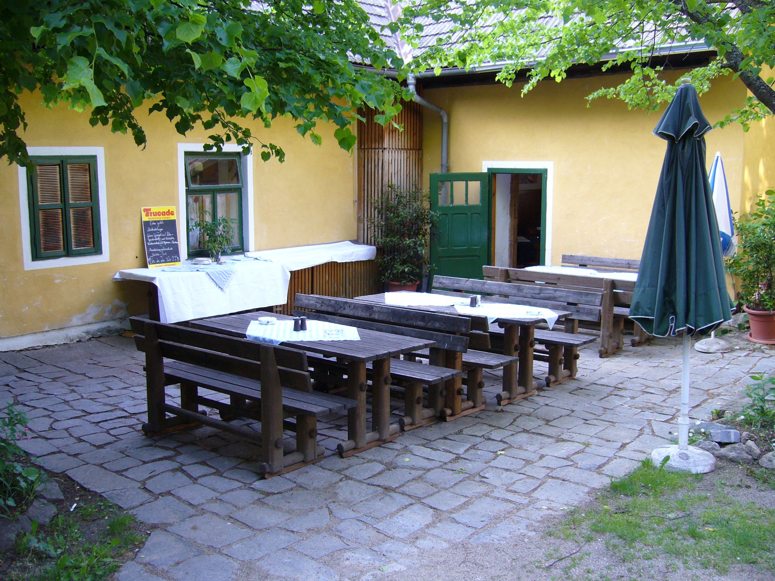 Inner courtyard of an inn with wooden benches and tables, yellow wall, green door and window, parasol and plants.