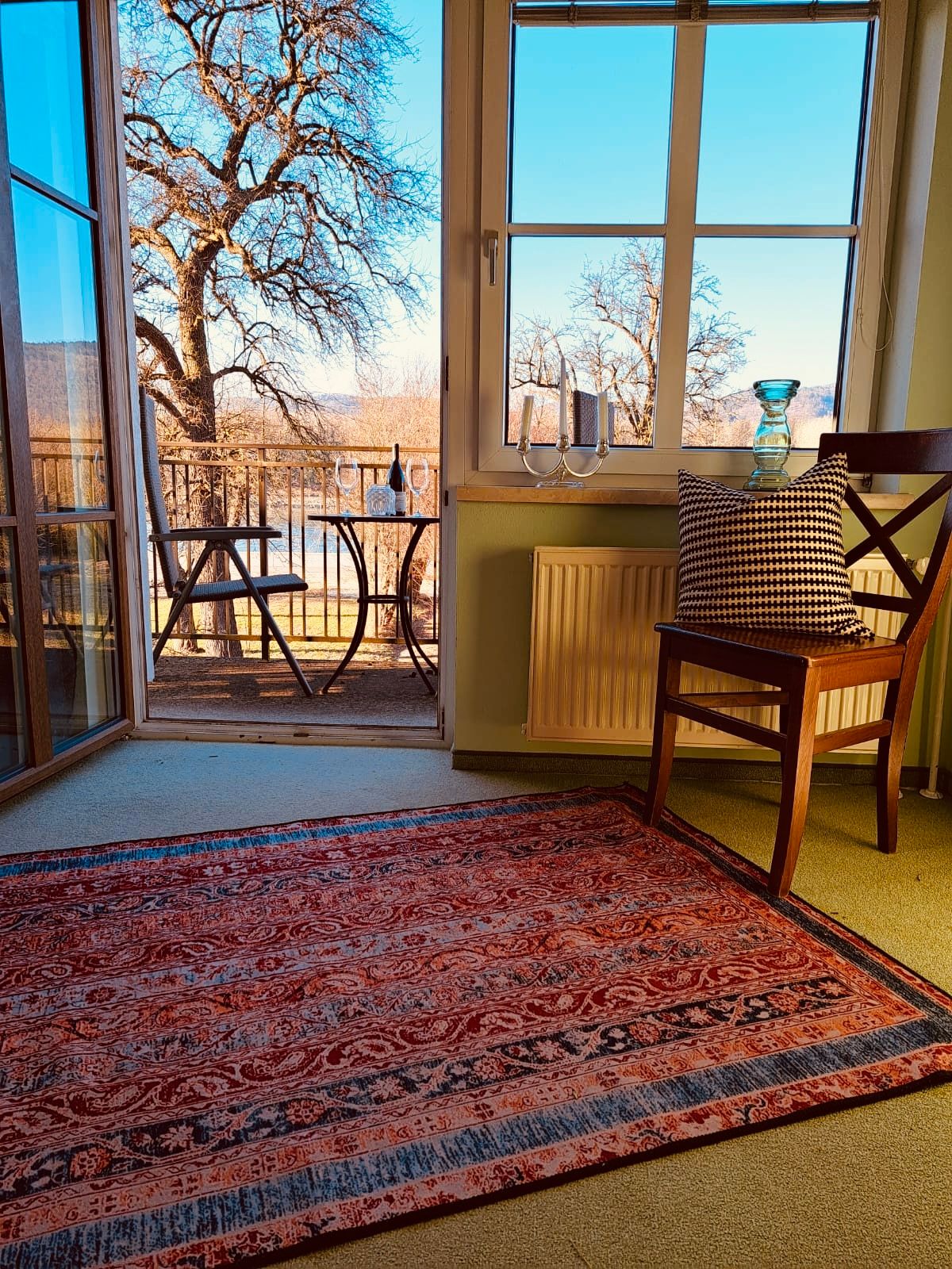 A room with carpet, chair and view of a balcony with table and chairs, trees in the background.