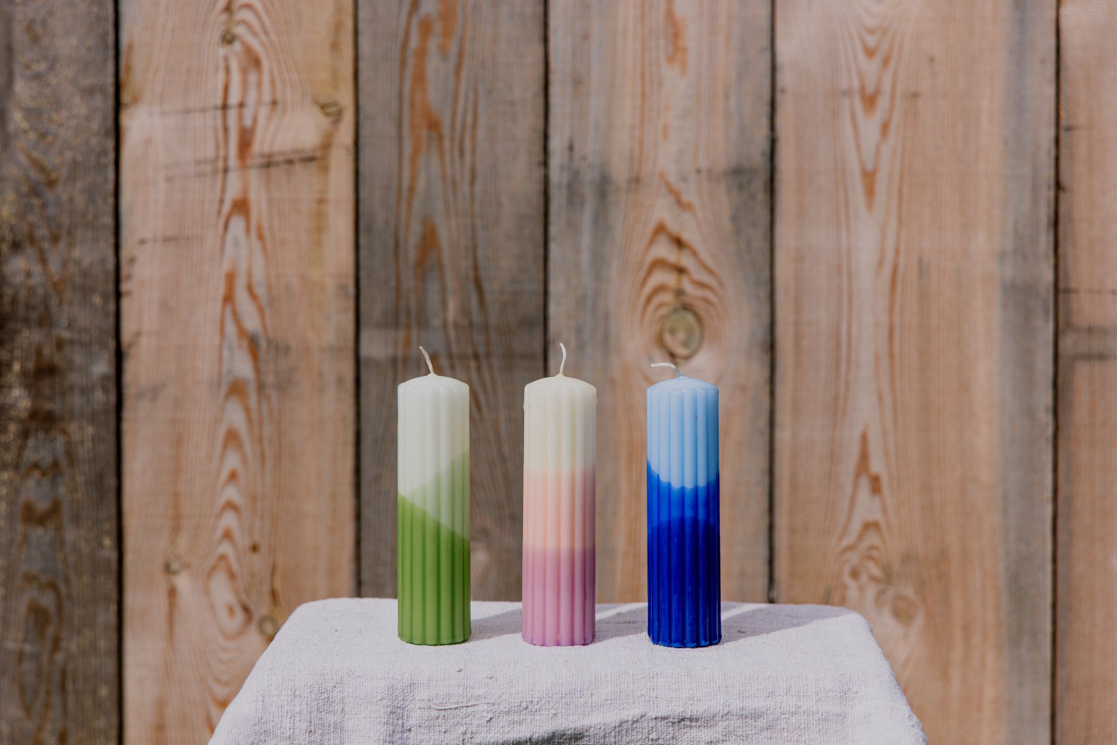 Three colorful candles on a table in front of a wooden wall.