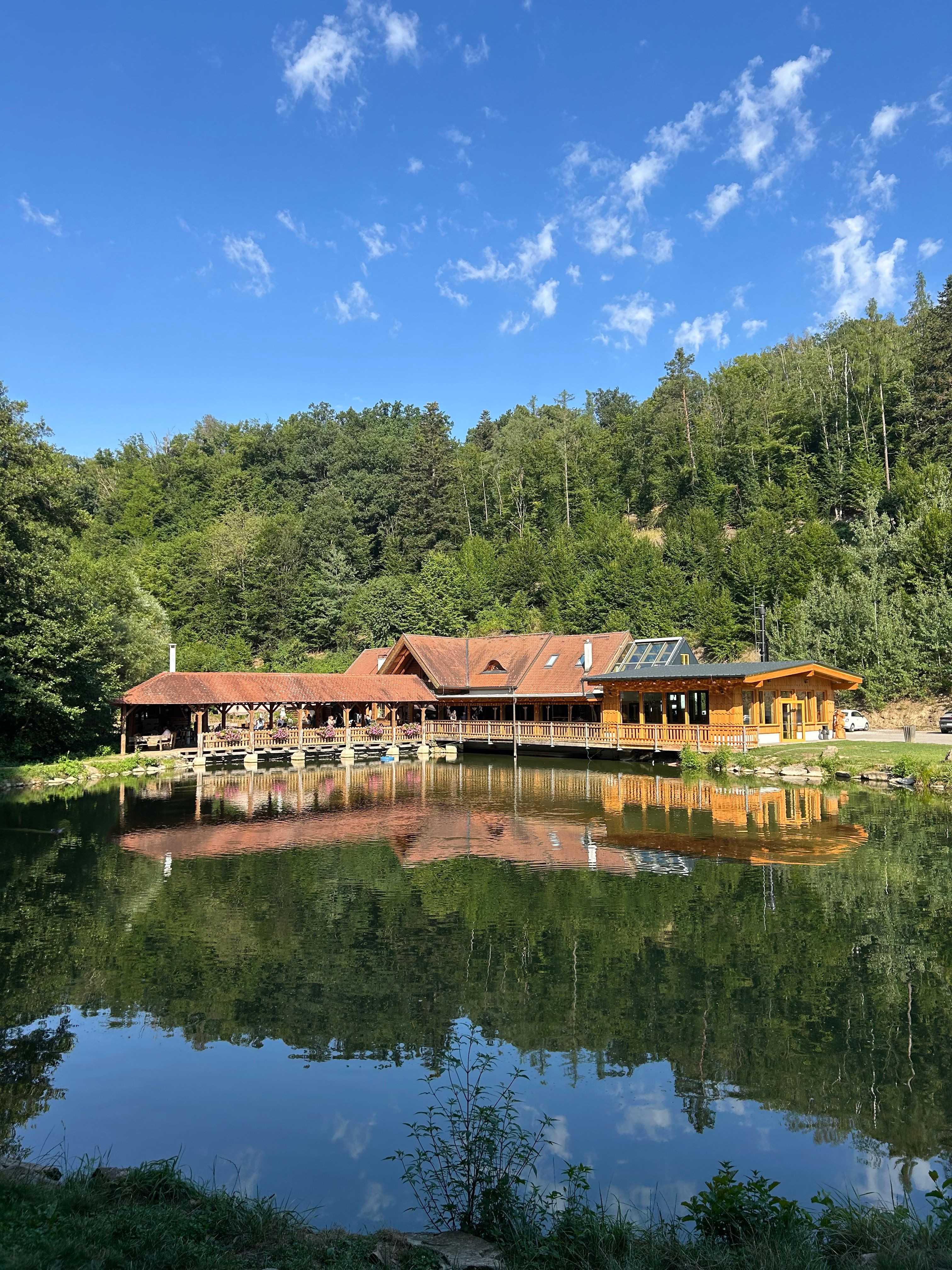 A wooden building on the shore of a lake, surrounded by woods, is reflected in the water. The sky is blue with a few clouds.