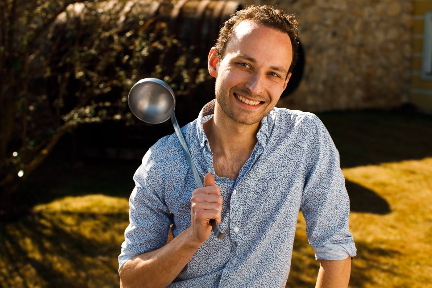 A smiling man outdoors holds a large ladle in his hand.
