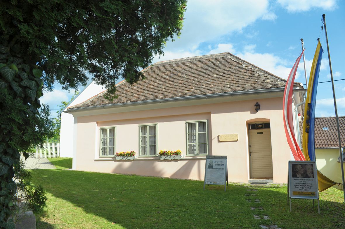 A small, pink-painted building with a shingle roof, surrounded by trees and flags, with signs in front of the entrance.