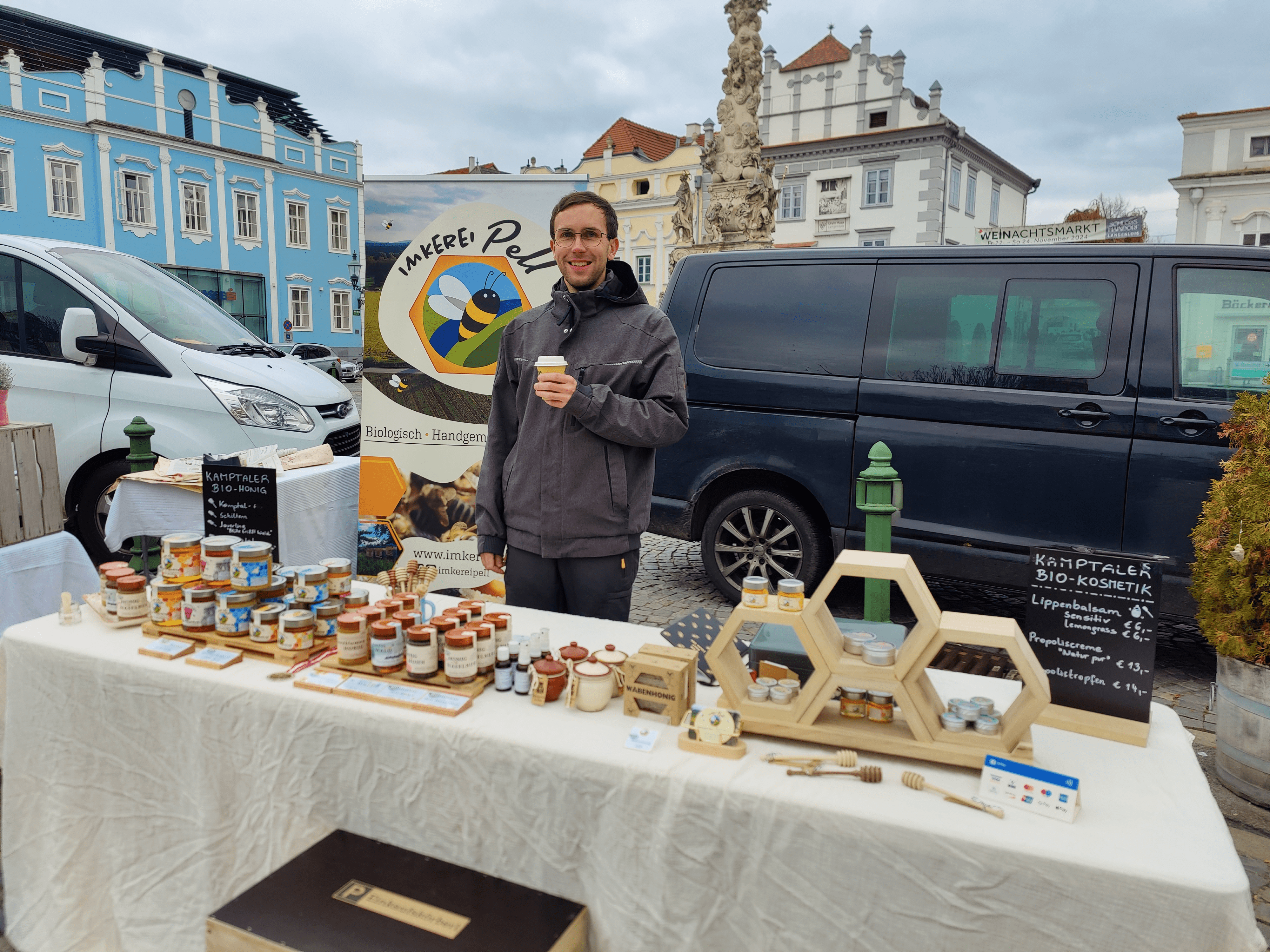 A man stands at a market stall with honey products and holds a cup. A van and buildings can be seen in the background.