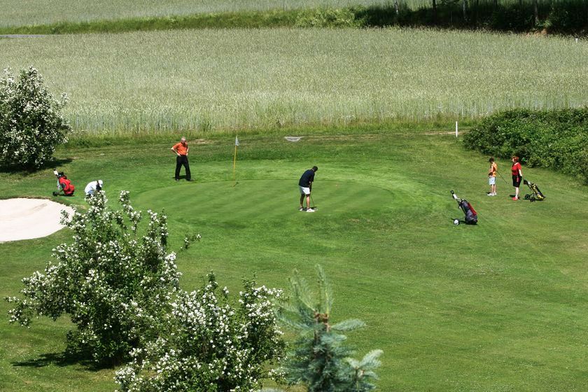 People play golf on a green course with trees and a sand bunker.