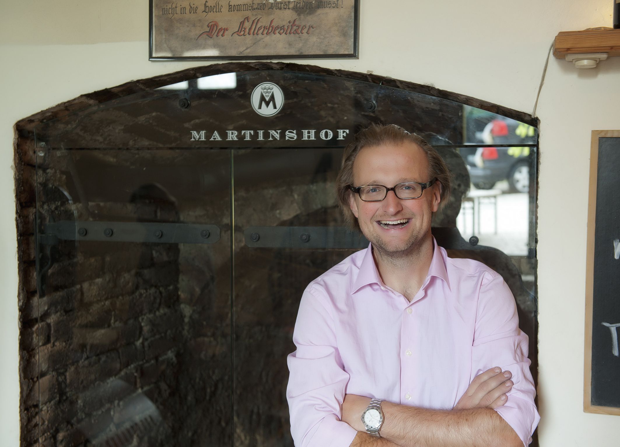 A man with glasses and a pink shirt stands smiling in front of a sign that reads 'Martinshof'.
