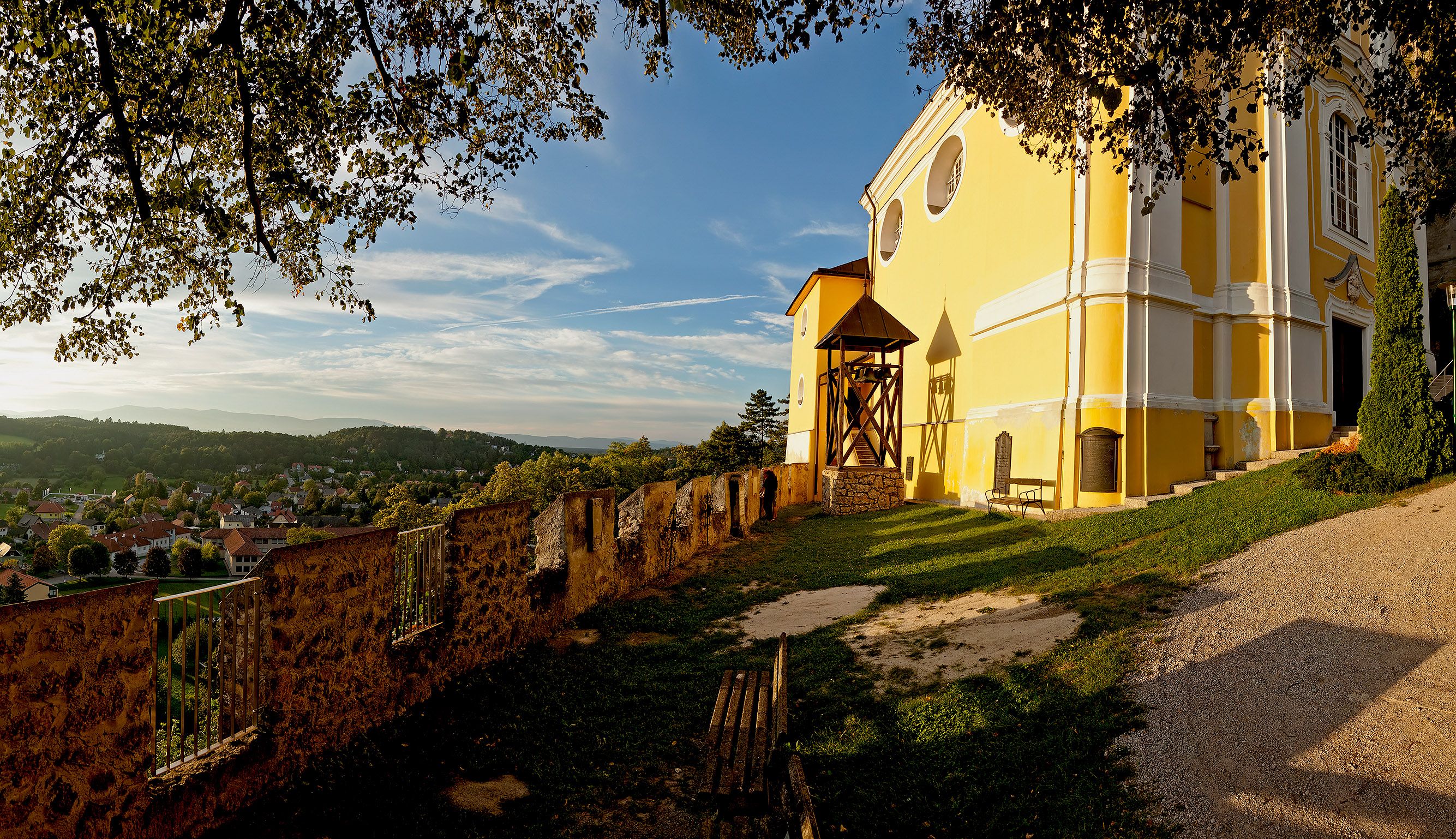 Yellow church on a hill with a view of a village and wooded hills in the background.