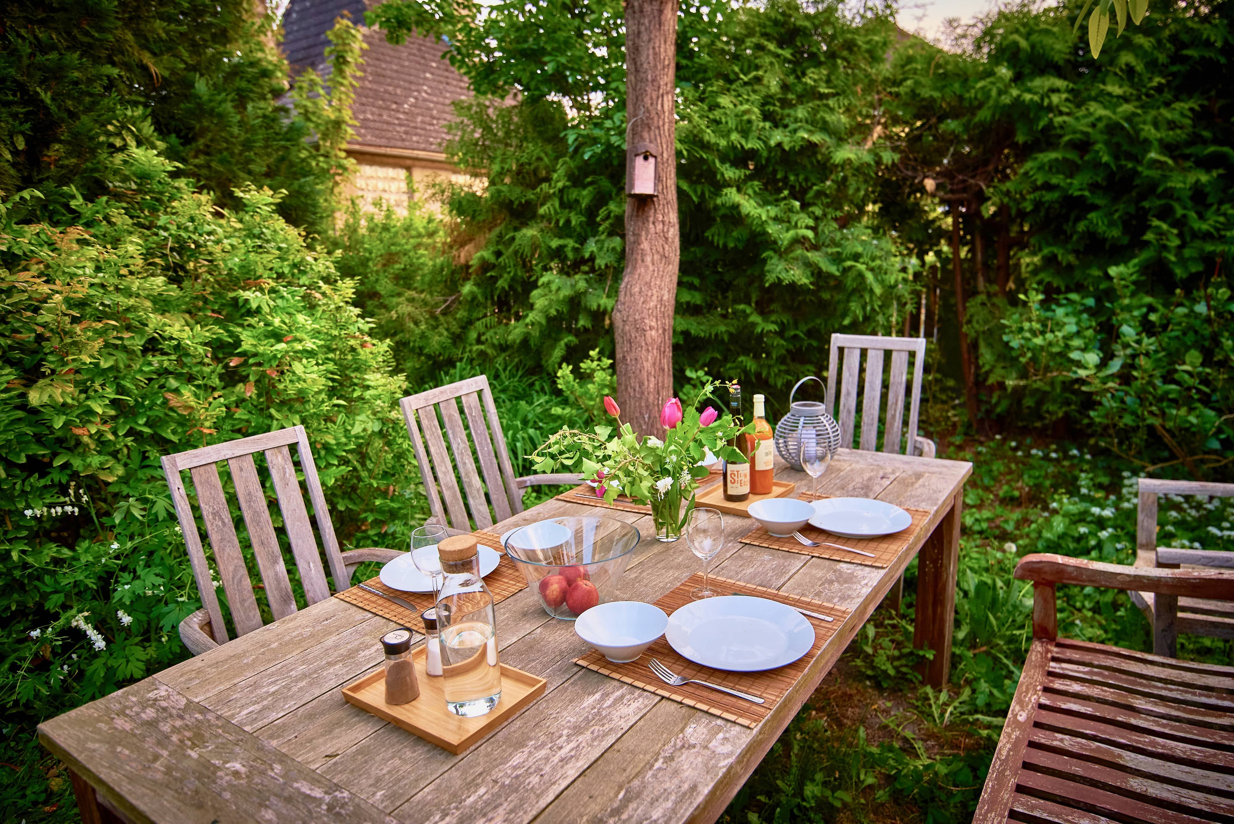 A wooden table set in the garden with plates, glasses, a vase of flowers and a bottle of wine.