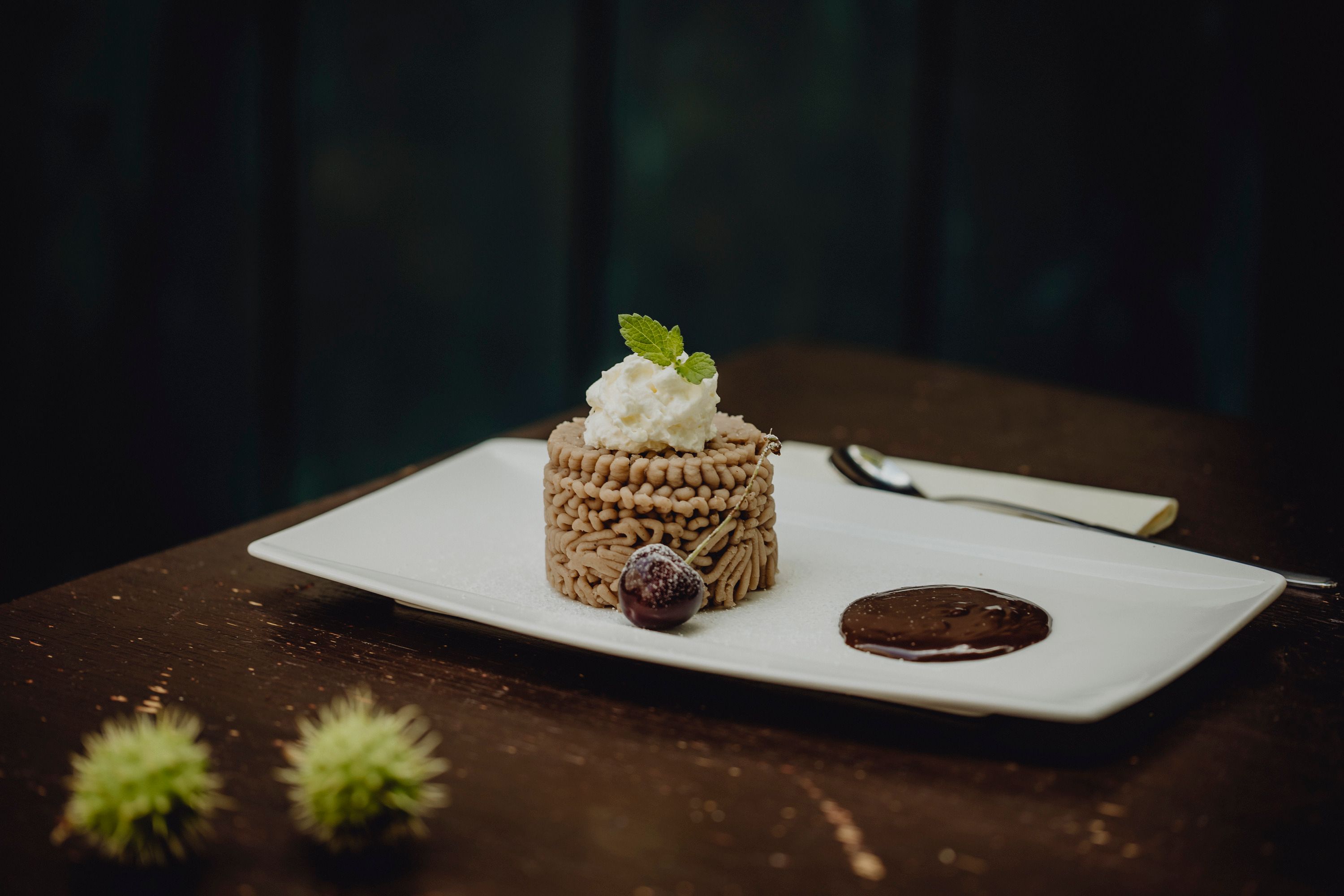 A dessert of chestnut mousse on a white plate, garnished with cream and a mint leaf, next to a chocolate sauce and a cherry.