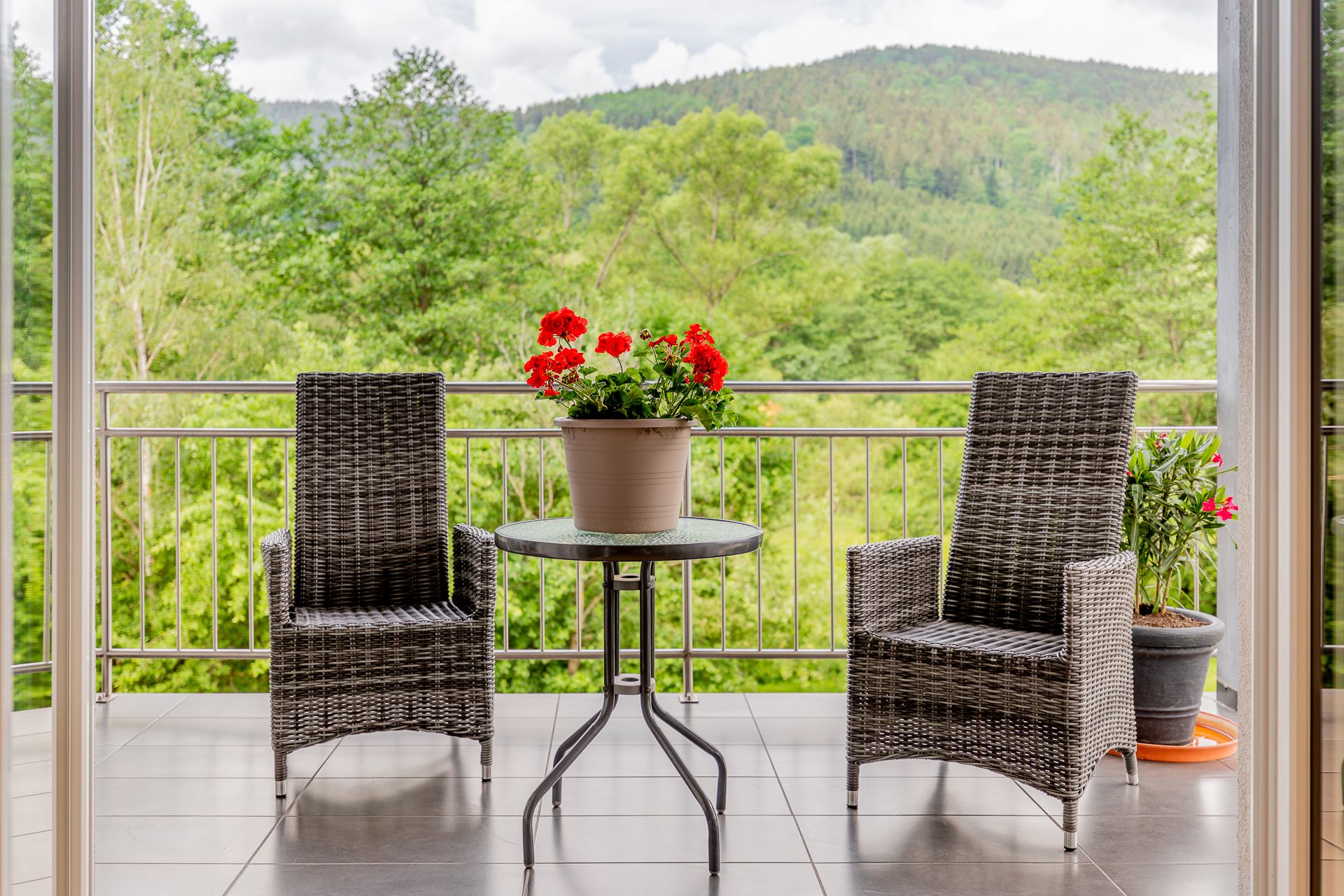 Balcony with two rattan chairs, a table with a flower pot and a view of wooded hills.