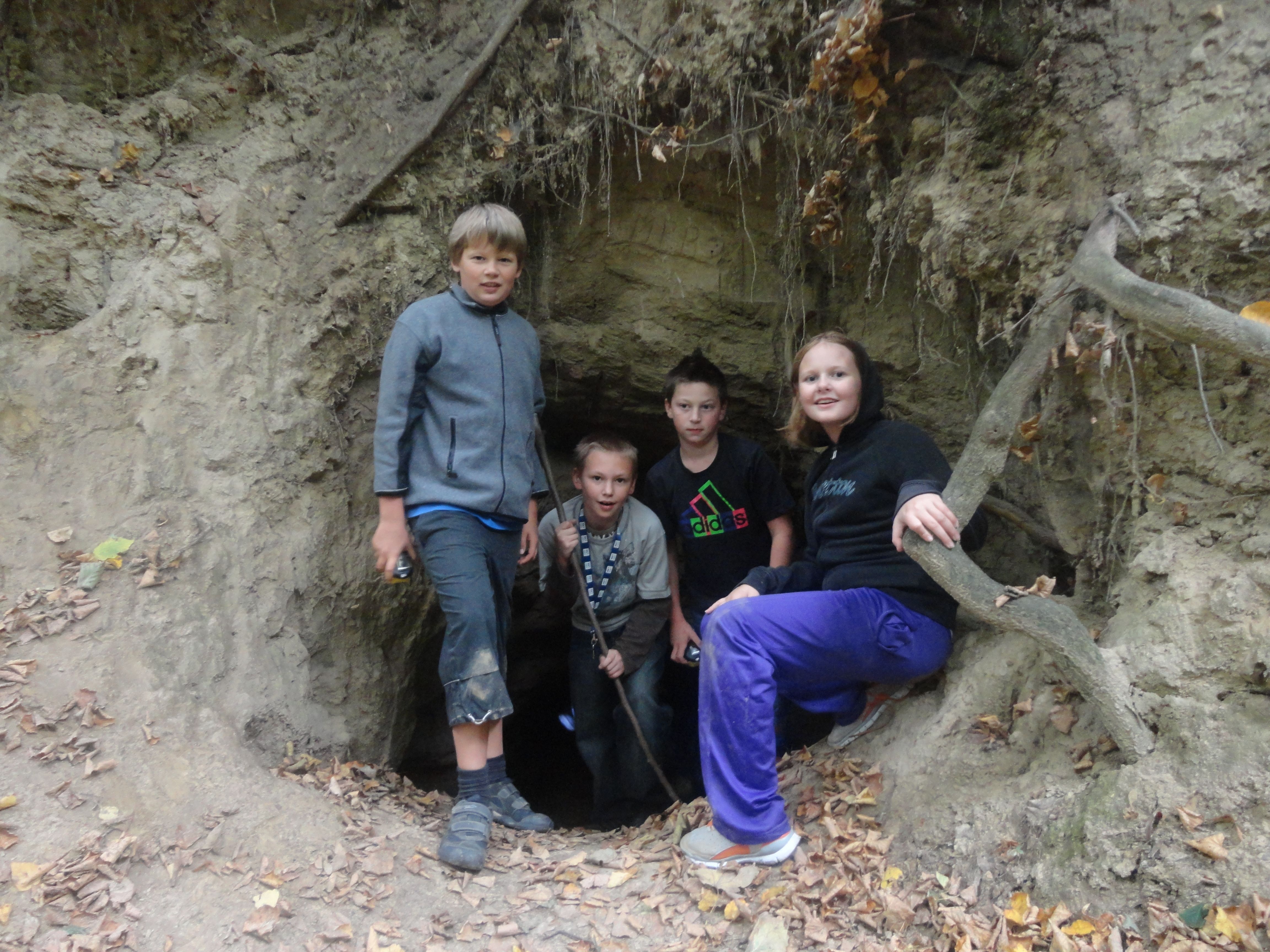 Four children stand at the entrance to a small cave in the forest.
