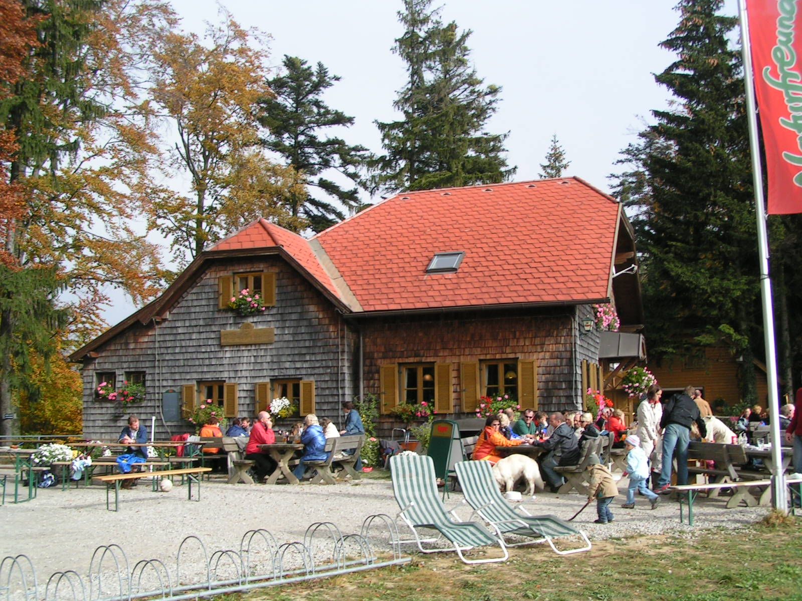 A mountain hut with a red roof and wooden walls, surrounded by trees. People are sitting at tables outside, some with dogs.