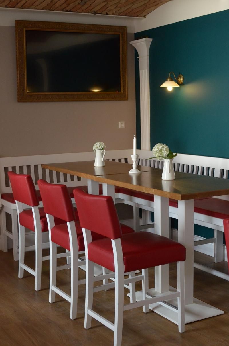 A dining area with red chairs, a wooden table, white vases and a candle. On the wall, a large, empty picture frame and a lamp.