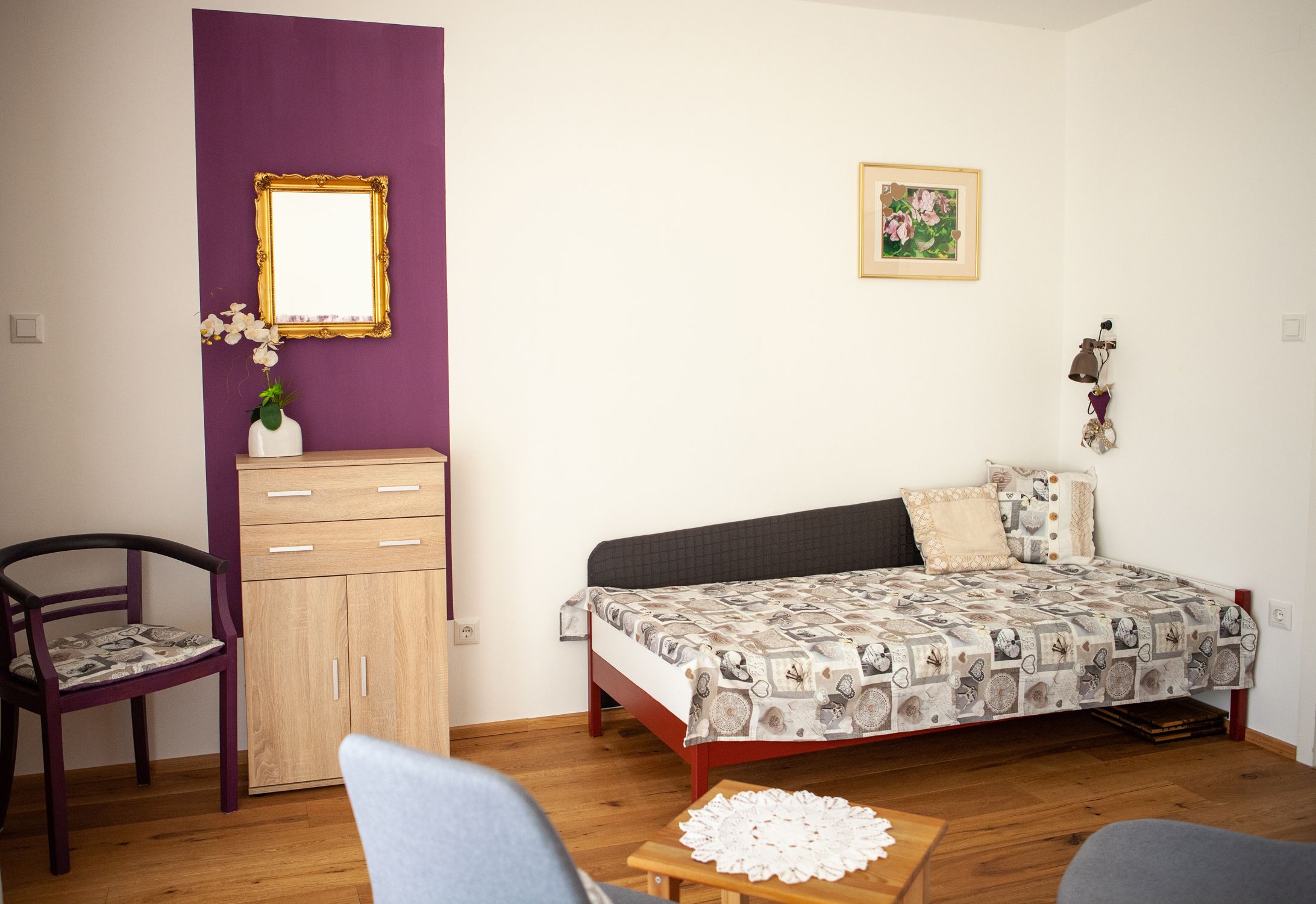 A living room with a sofa bed, a small wardrobe, a chair and a table on a wooden floor.