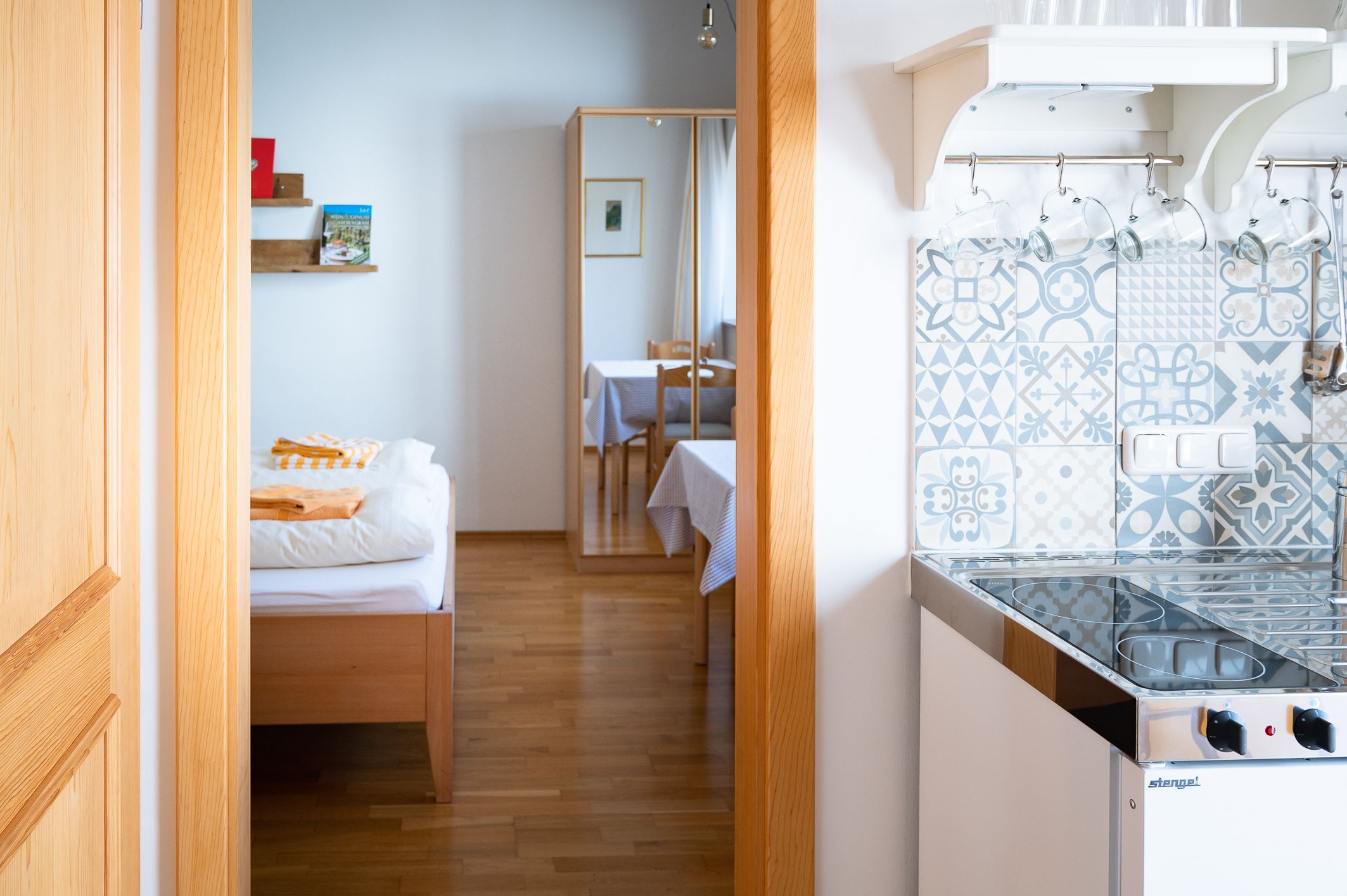 Interior view of a room with bed, mirror, table and small kitchen with patterned tiles.