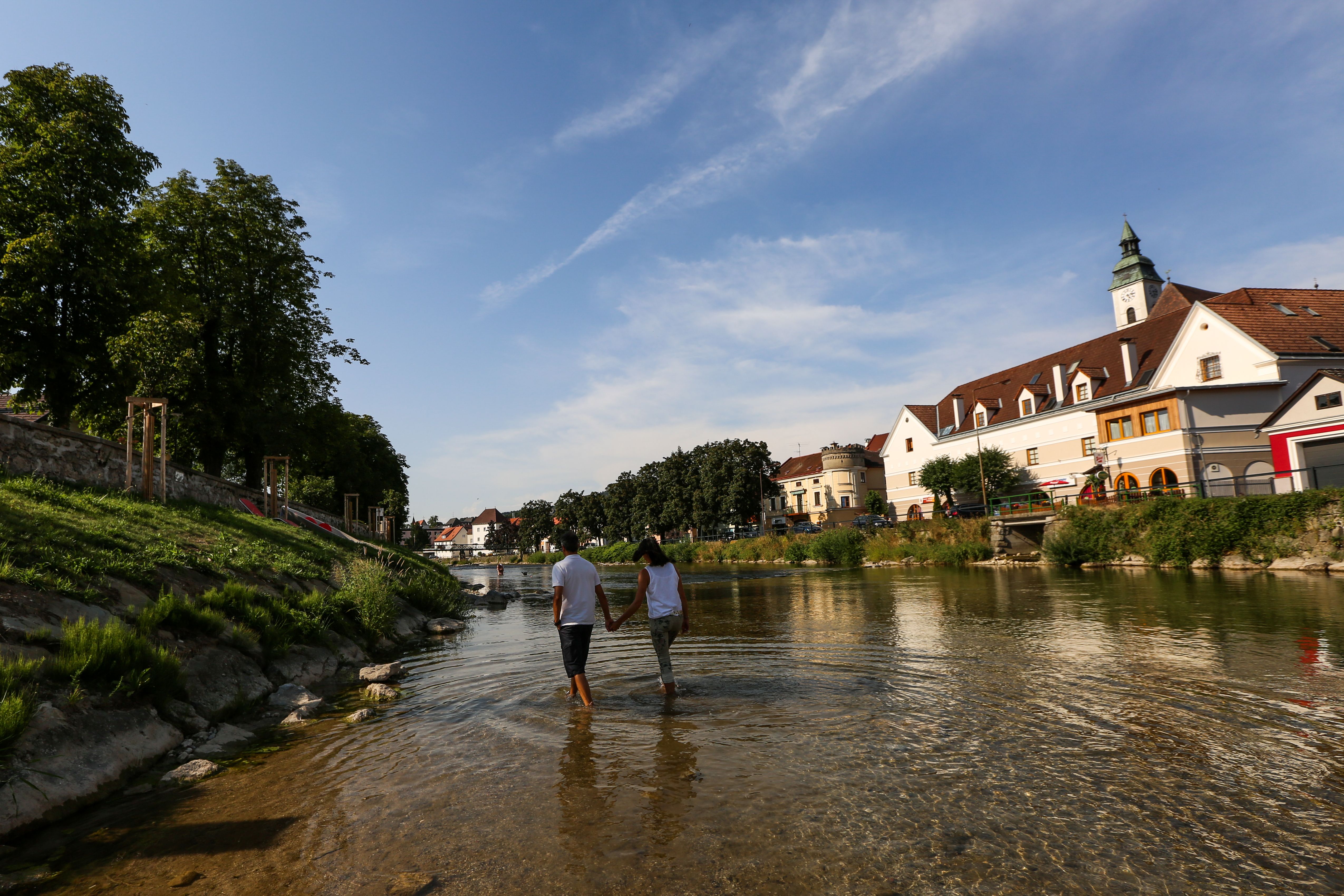 A couple walks hand in hand through a shallow river in a town with historic buildings and trees.