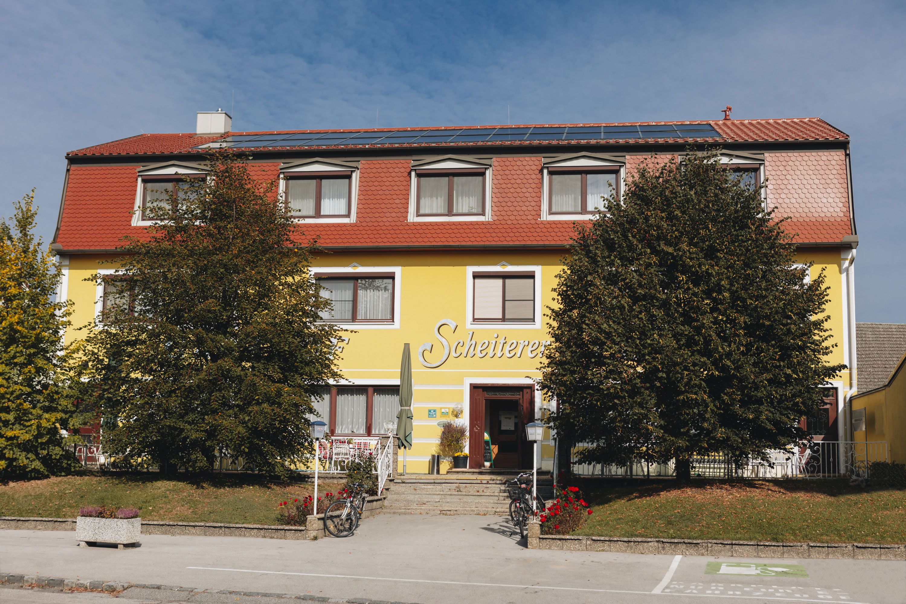 Yellow building with a red roof and the inscription 'Scheiterer', surrounded by trees and bicycles.