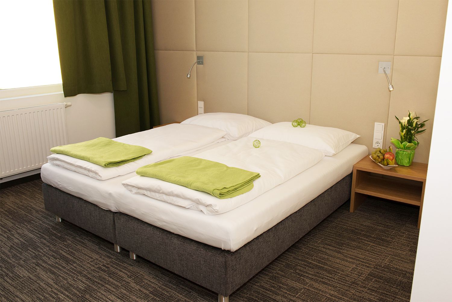 Comfortable hotel room with double bed, green towels and bedside table with flowers and fruit.