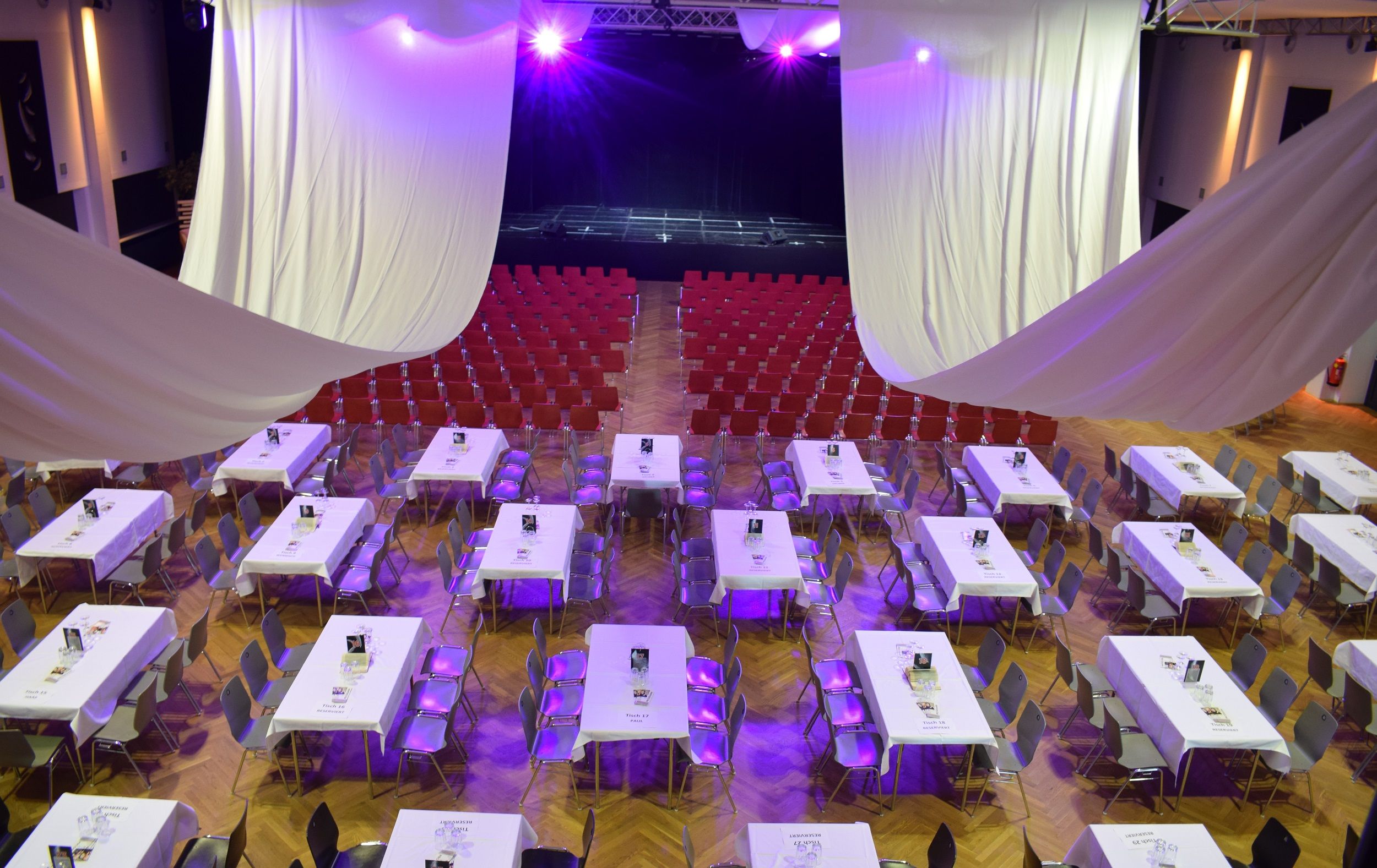 An event room with tables and chairs, purple lighting and a stage in the background.