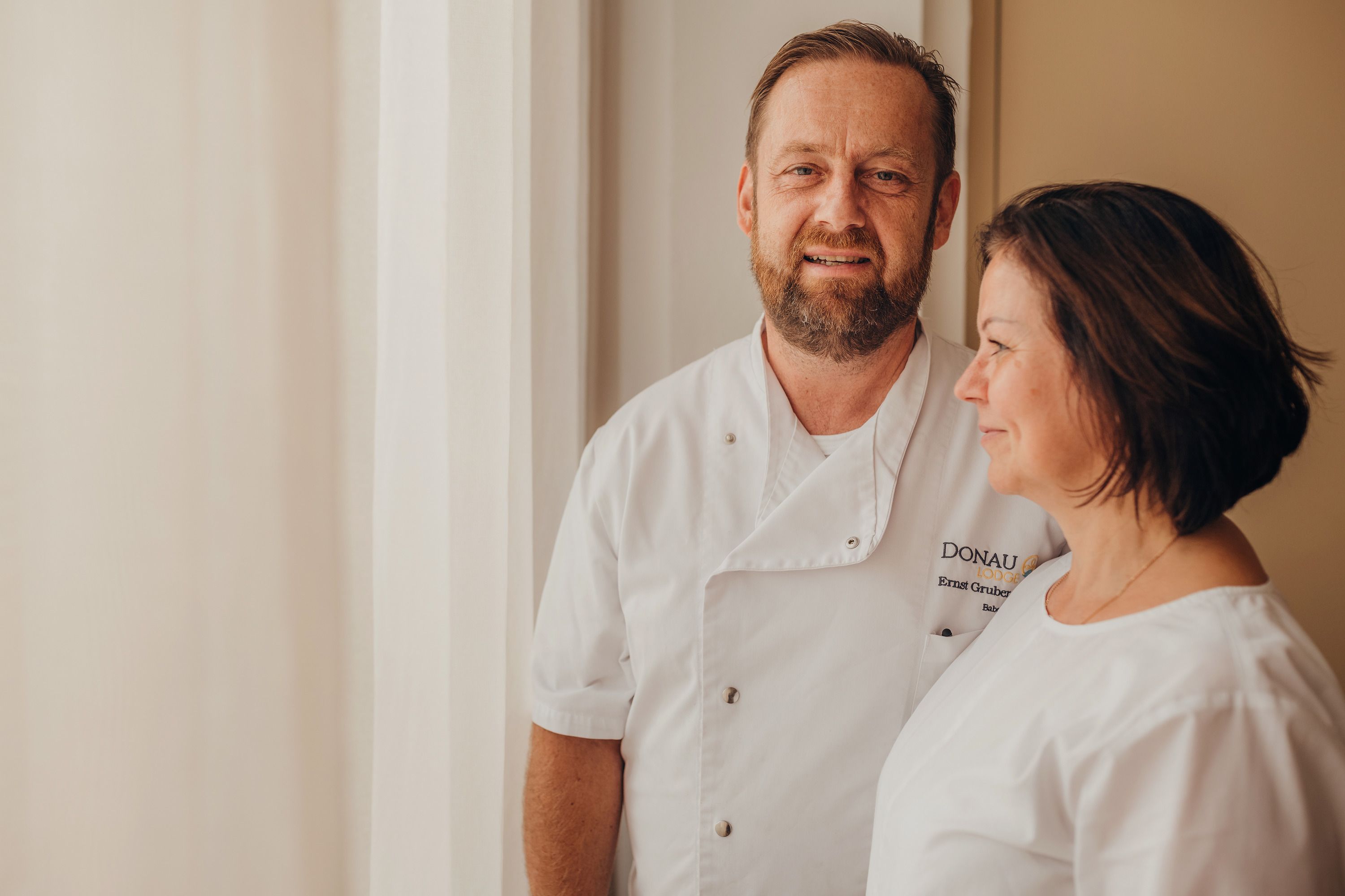 A man and a woman in white chef's jackets stand next to each other in front of a window.
