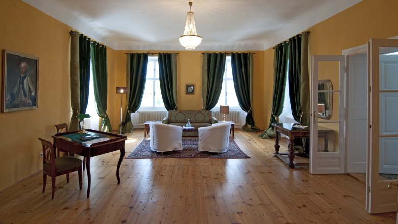 Elegant room with wooden floor, green curtains, two armchairs, a sofa and a desk. A chandelier hangs from the ceiling.