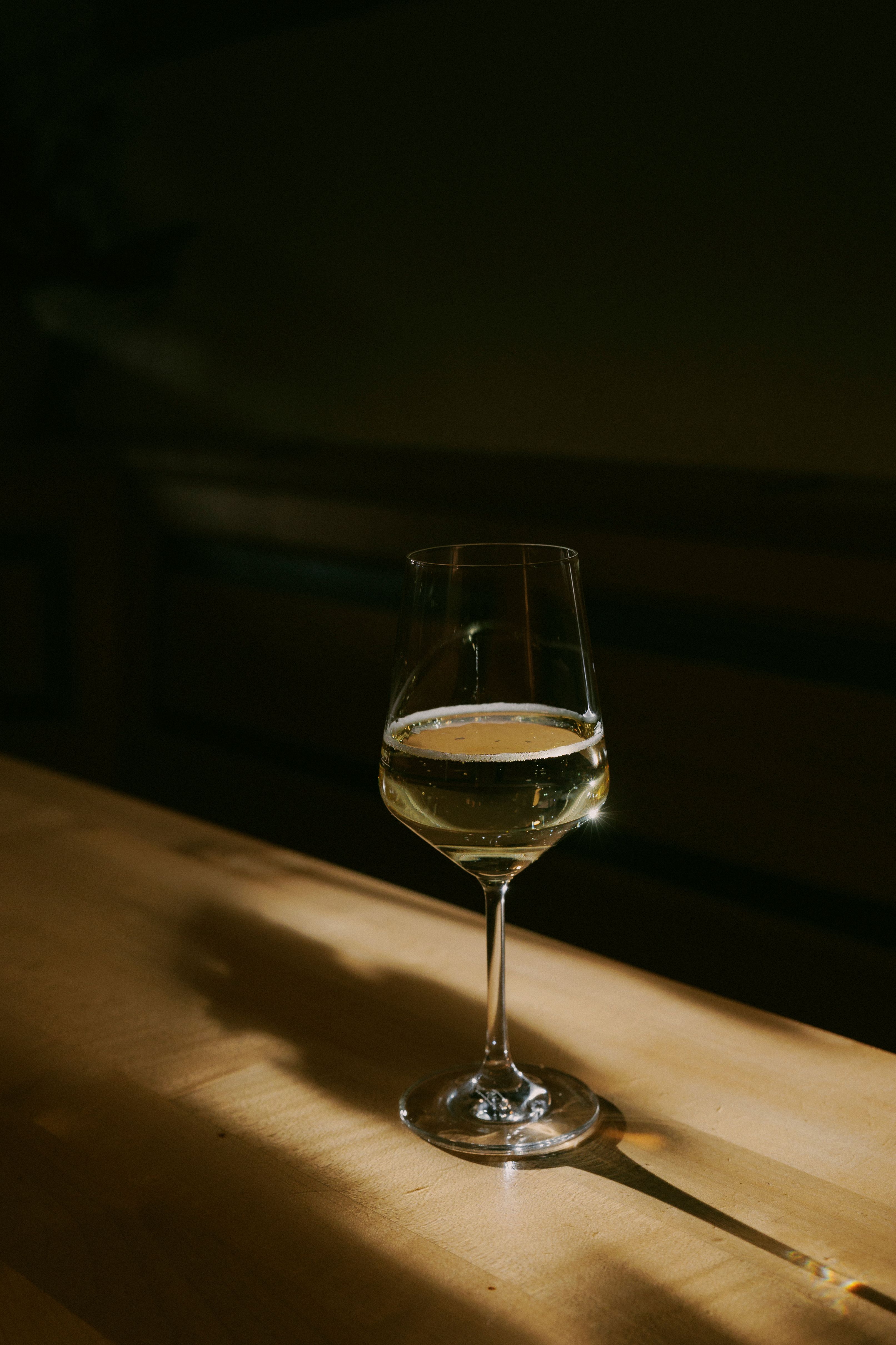 A wine glass with white wine stands on a wooden table in the shade.