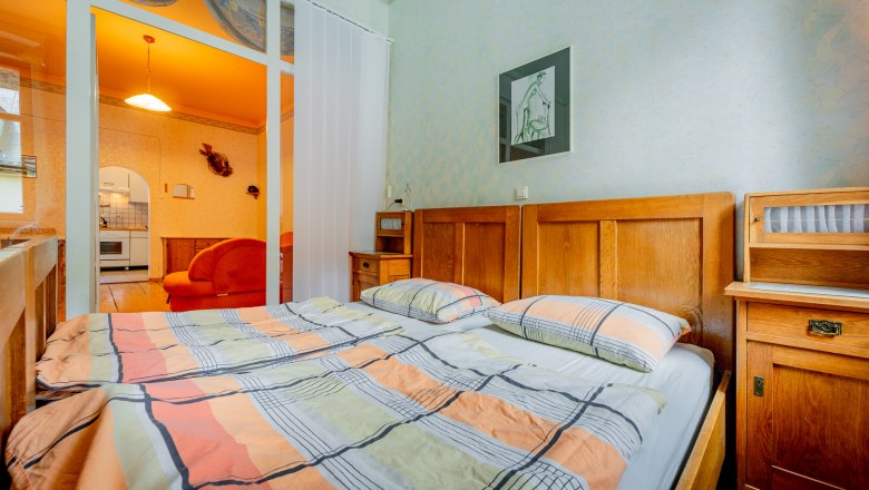 Bedroom with double bed, wooden furniture and view into a living room with orange lighting.