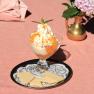 A dessert glass with ice cream, whipped cream and apricots on a tray with waffles, next to a vase with flowers.