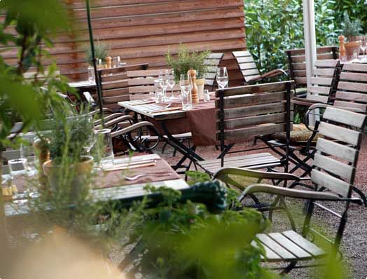 A cozy guest garden with wooden tables and chairs, set with glasses and cutlery, surrounded by plants.
