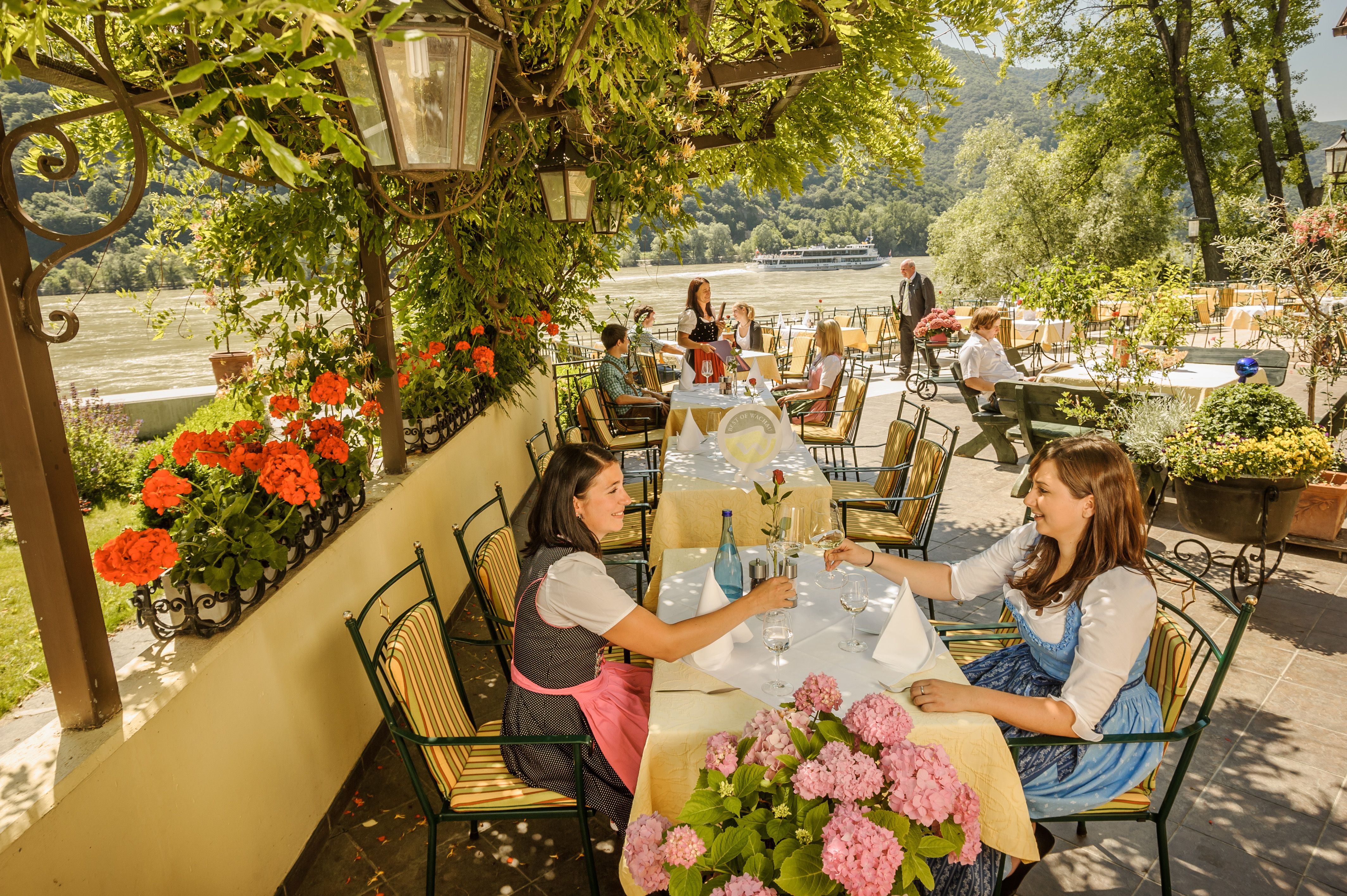People sitting in a garden restaurant with a view of a river, surrounded by flowers and trees.