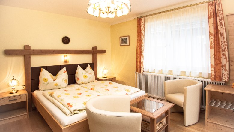 Cozy bedroom with double bed, two armchairs and a table. Bright curtains and warm lighting.