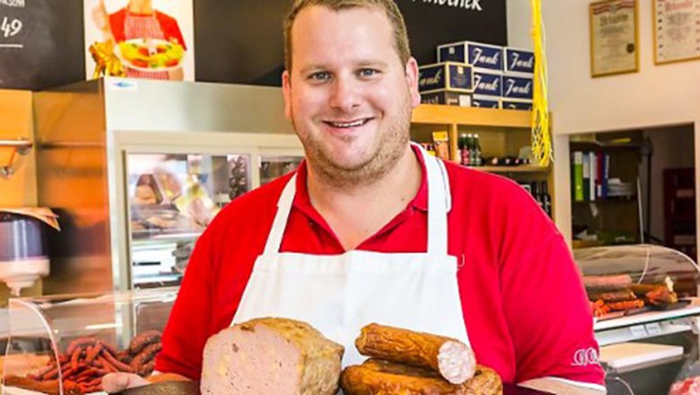 A butcher in a red shirt and white apron holds various sausage products in a butcher's shop.