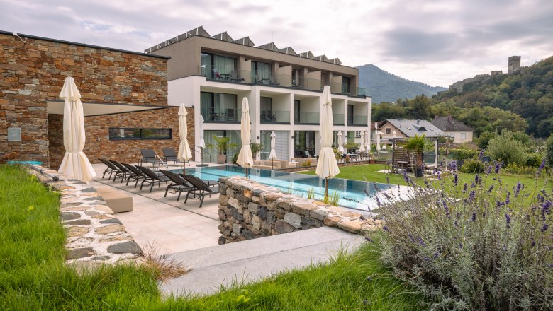 Exterior view of a modern boutique hotel with pool and sun loungers, surrounded by green countryside and hills.