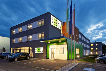 Modern building with the inscription 'Good Rooms' at dusk, surrounded by cars and flags.