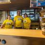Three people in yellow T-shirts are standing behind a bar in a rustic room.