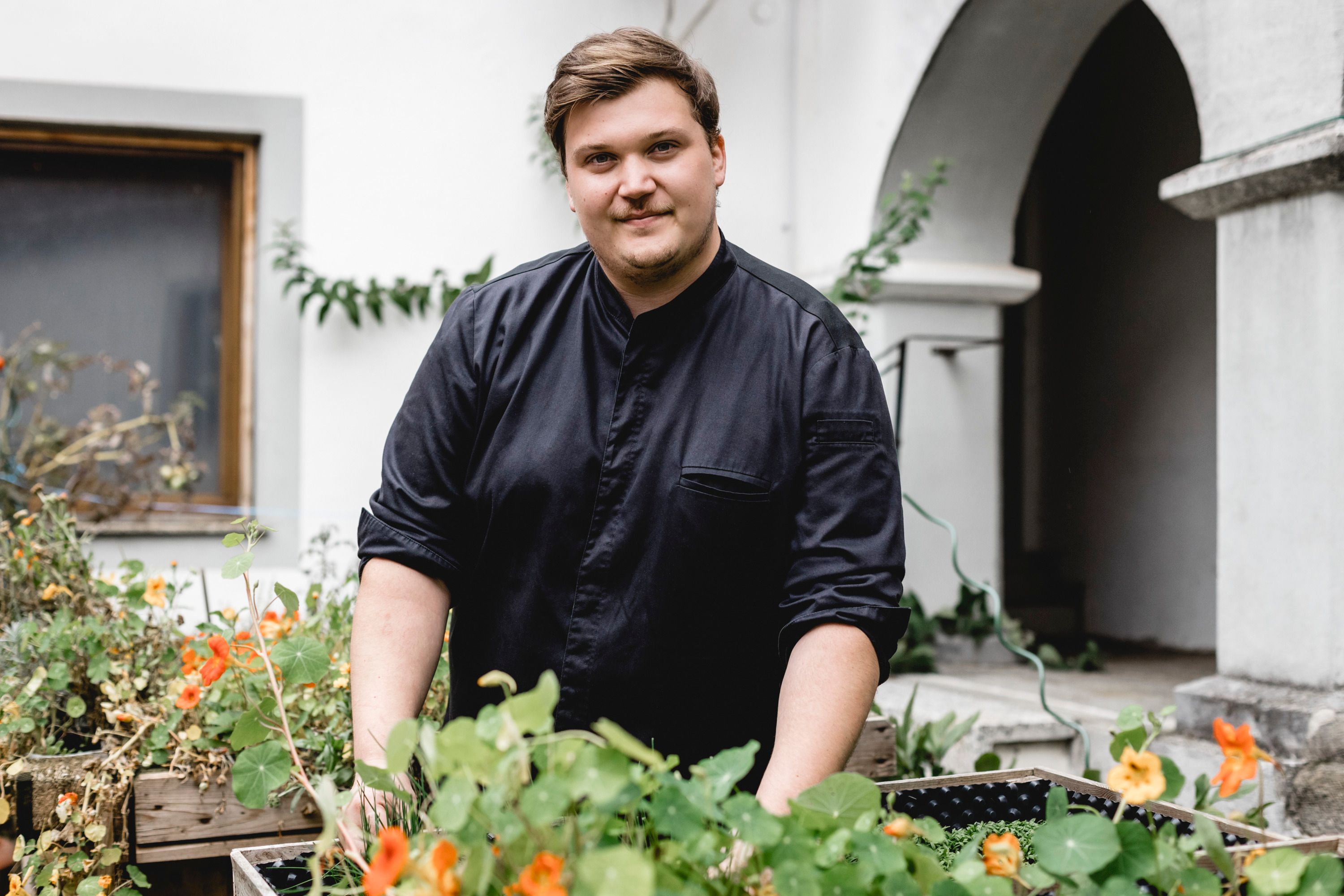 A man in black clothing stands in a garden with flowers and plants.
