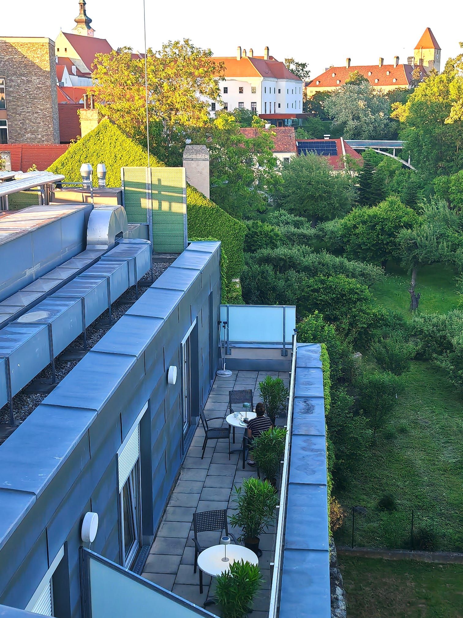 A view from above of a rooftop terrace with tables and chairs, surrounded by green plants. In the background, you can see buildings with red roofs and trees.
