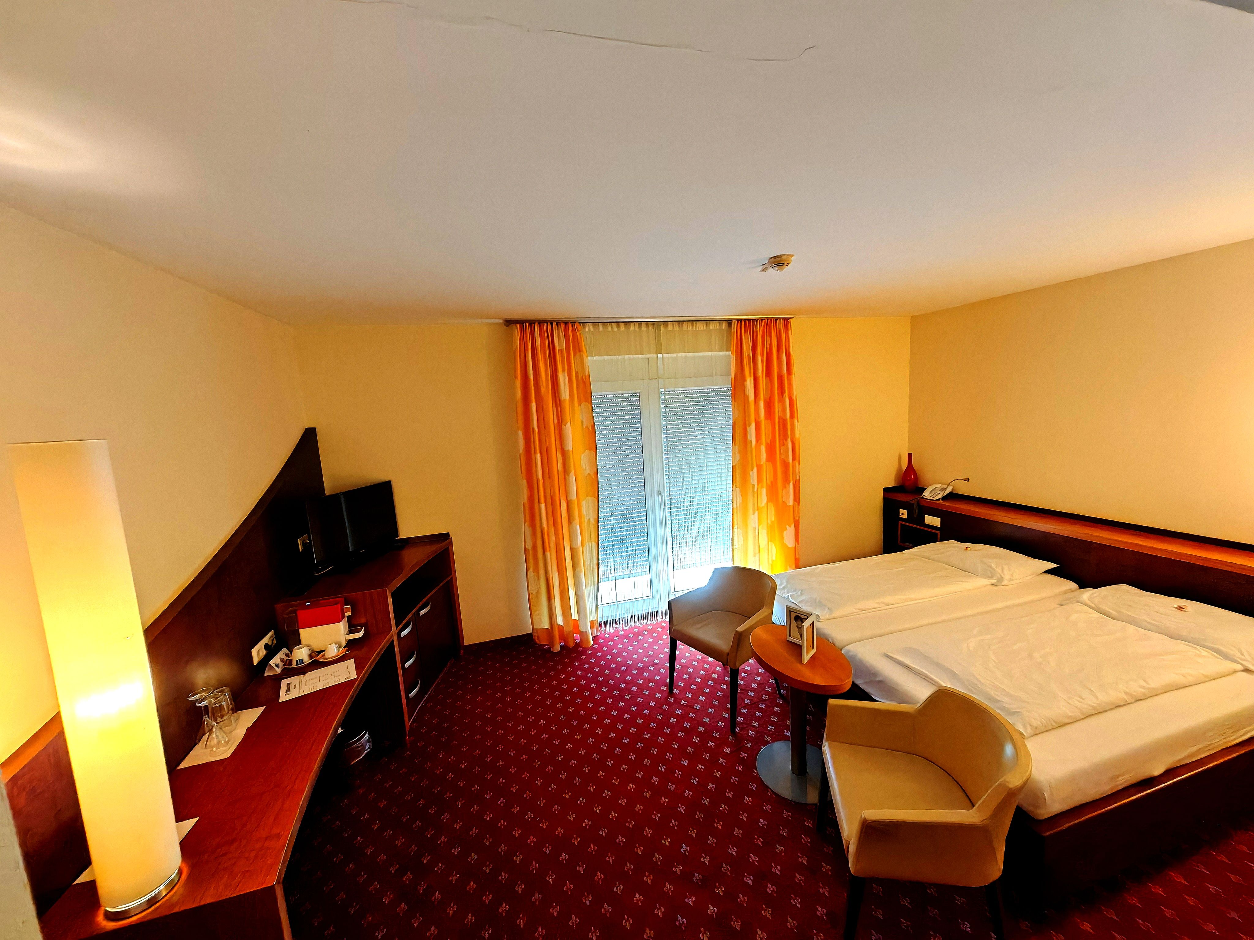 A hotel room with a double bed, two chairs, a table, a desk with a TV, and a floor lamp.