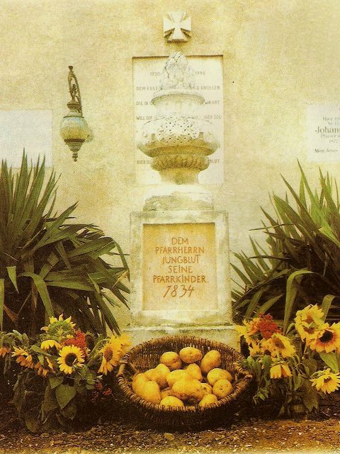 Monument with inscription, surrounded by plants and a basket of potatoes and sunflowers.